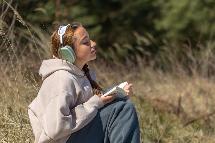 Nuori nainen kuuntelee musiikkia kuulokkeilla luonnossa kirja kädessään